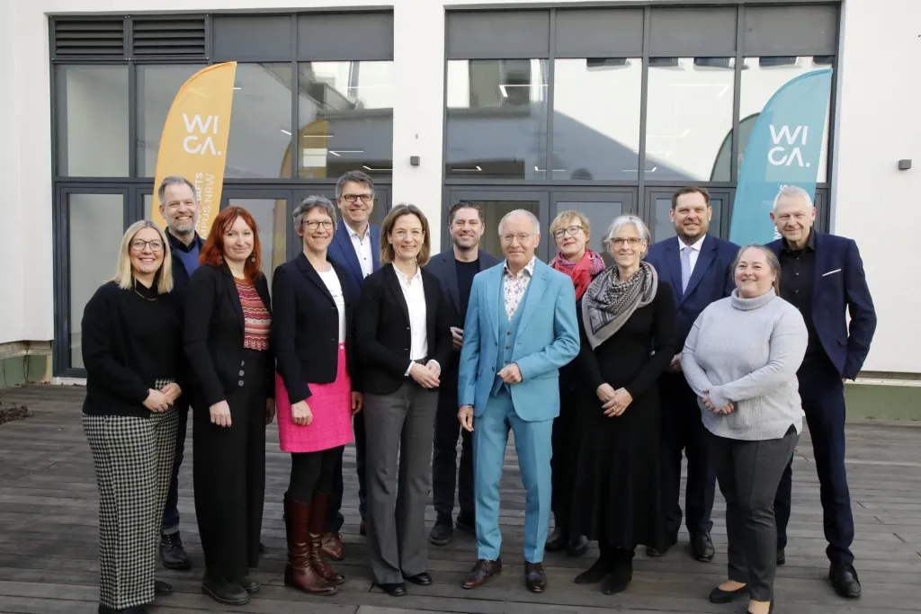 Gruppenfoto von dreizehn Personen vor WICA-Fahnen im Freien