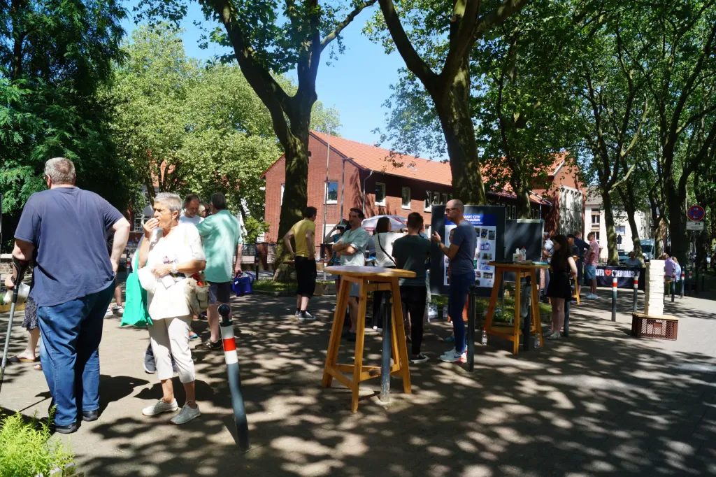 Straßenaktion mit Stehtischen und Infotafeln unter Bäumen, Menschen im Gespräch vor einem Backsteingebäude