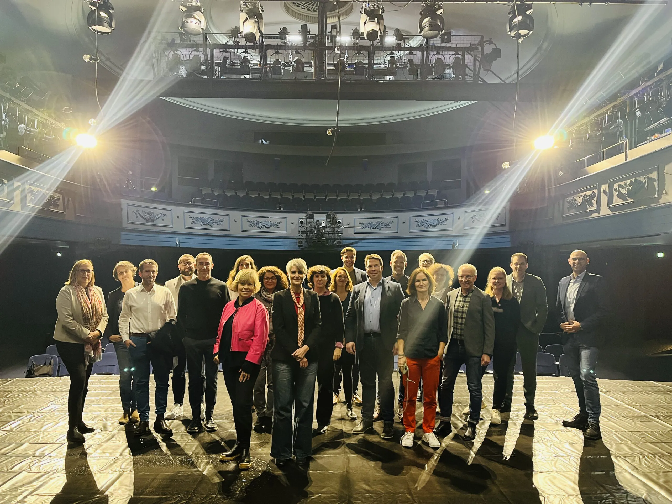Gruppenbild auf einer Theaterbühne, dahinter die Ränge des Zuschauerraums im Scheinwerferlicht