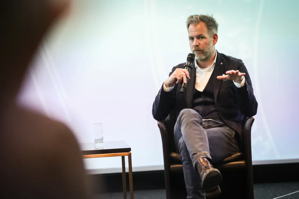 Mann mit Bart spricht gestikulierend mit Mikrofon in einem Sessel auf dem Podium