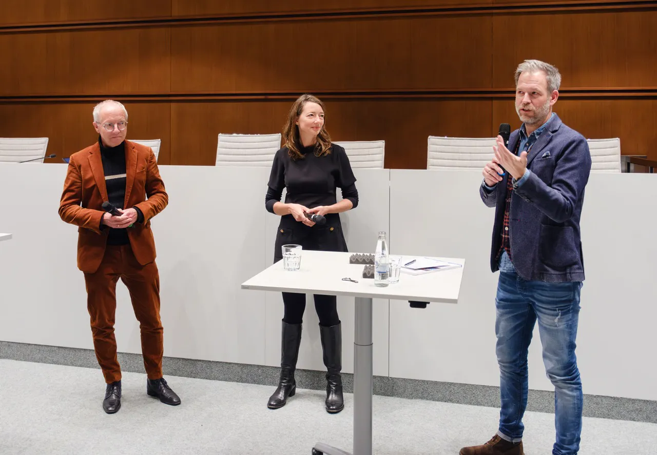 Drei Personen bei einer Gesprächsrunde im Ratssaal, ein Mann spricht ins Mikrofon